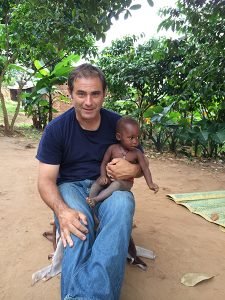 Dan Smith is holding a small child as he sits on the ground in an African village