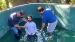 This is a picture of a man from India kneeling in a small swimming pool with two other men beside him as he gets baptized, taken during Hope City Supernatural Lifestyle Training missions trip to Inda.